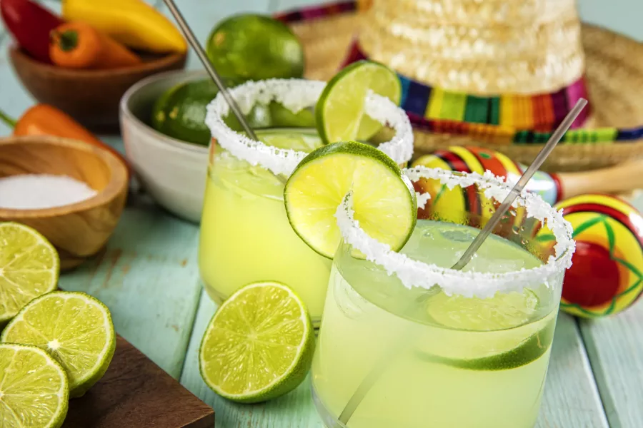 This is a photograph of two modern lime margarita glass with a rim of salt surrounded by fresh cut limes and chilis on a colorful Slated Wood Background next to a sombrero